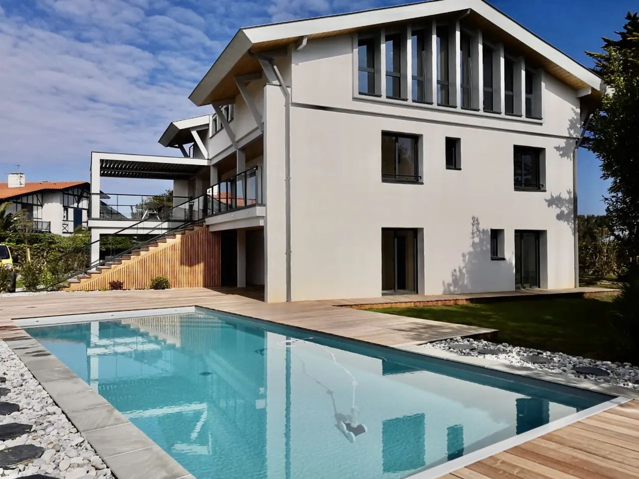 Piscine rectangulaire entourée d'une terrasse en bois et de galets blancs, devant une maison aux murs clairs